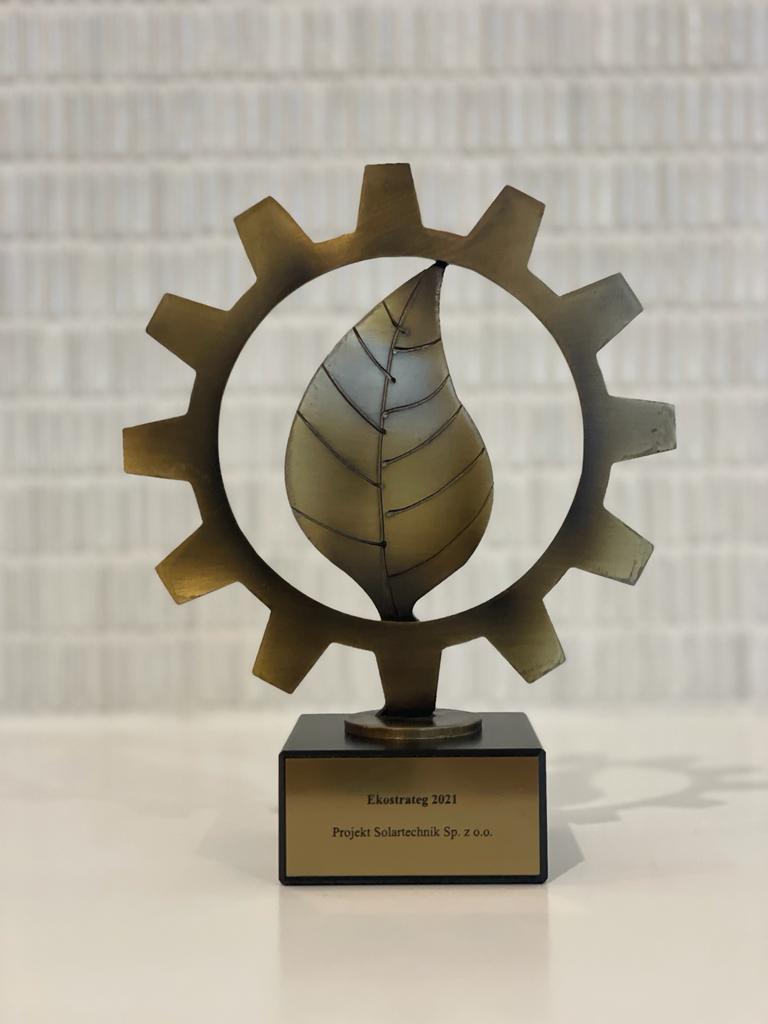 Złota statuetka przedstawiająca liść wpisany w koło zębate z tabliczką mówiącą ekostrateg 2021 Projekt Solartechnik na jasnym tle